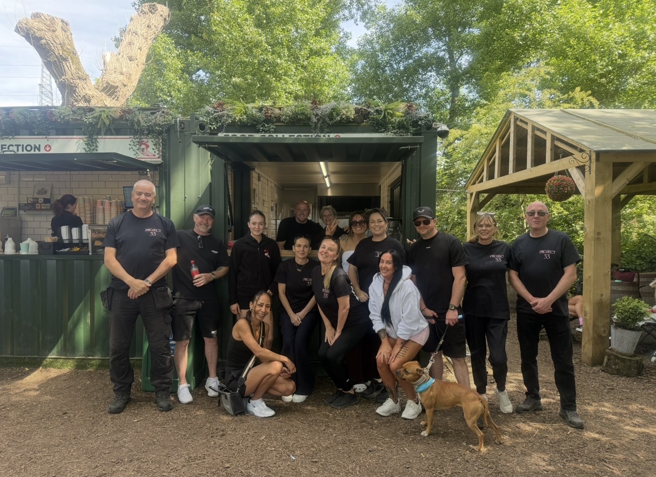 The Project 33 team and volunteers gathered together outdoors in front of a green community kiosk