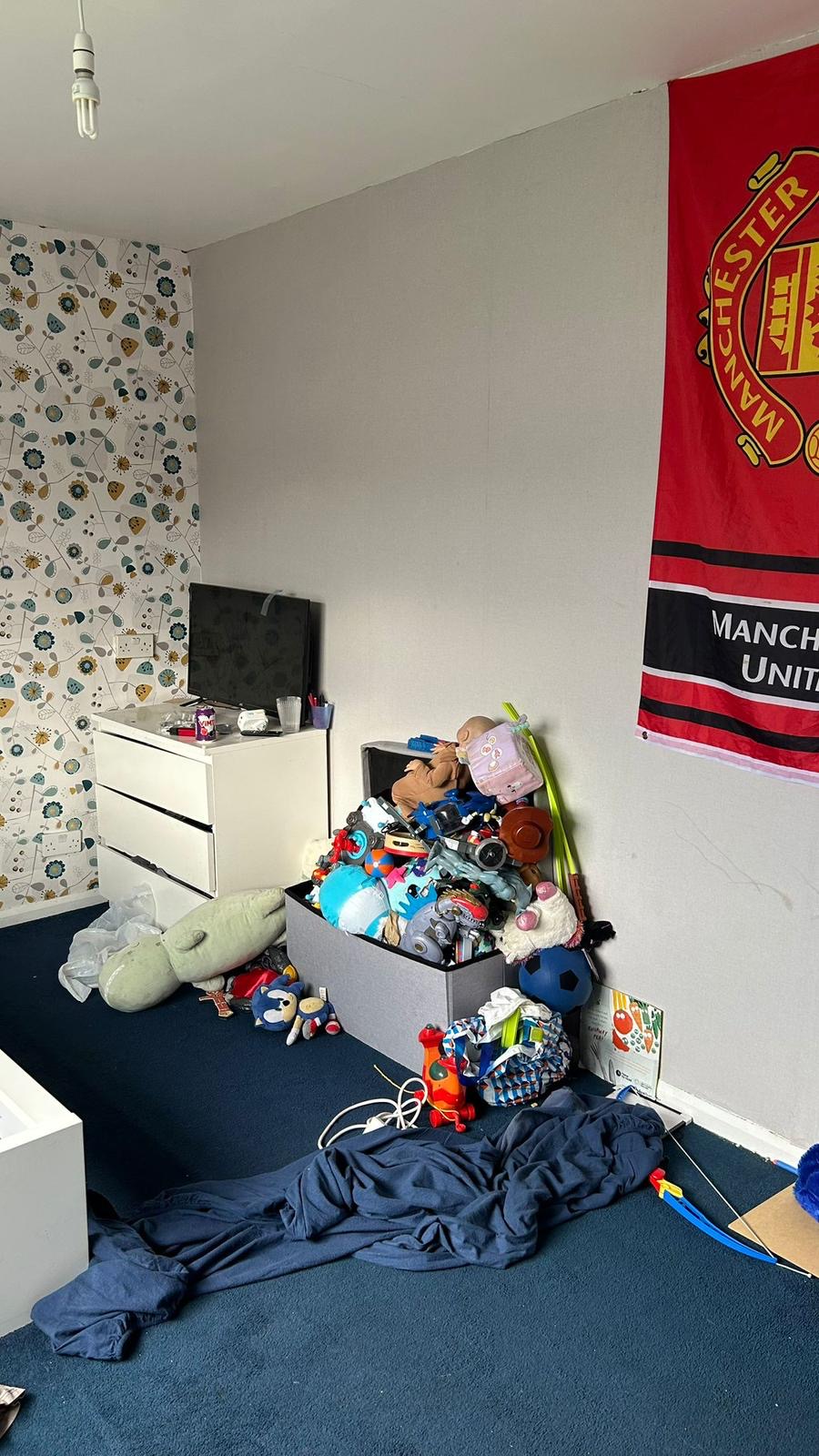 The boys' bedroom before: scattered toys, blue carpet covered with clothes, and a Manchester United flag on the wall