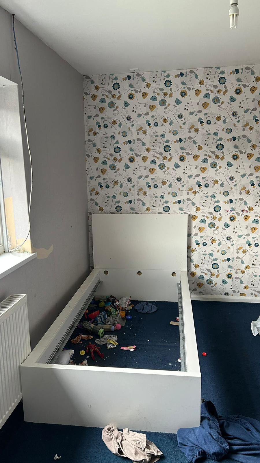 A child's bedroom before: stripped wallpaper, dismantled bed frame, clutter on the floor